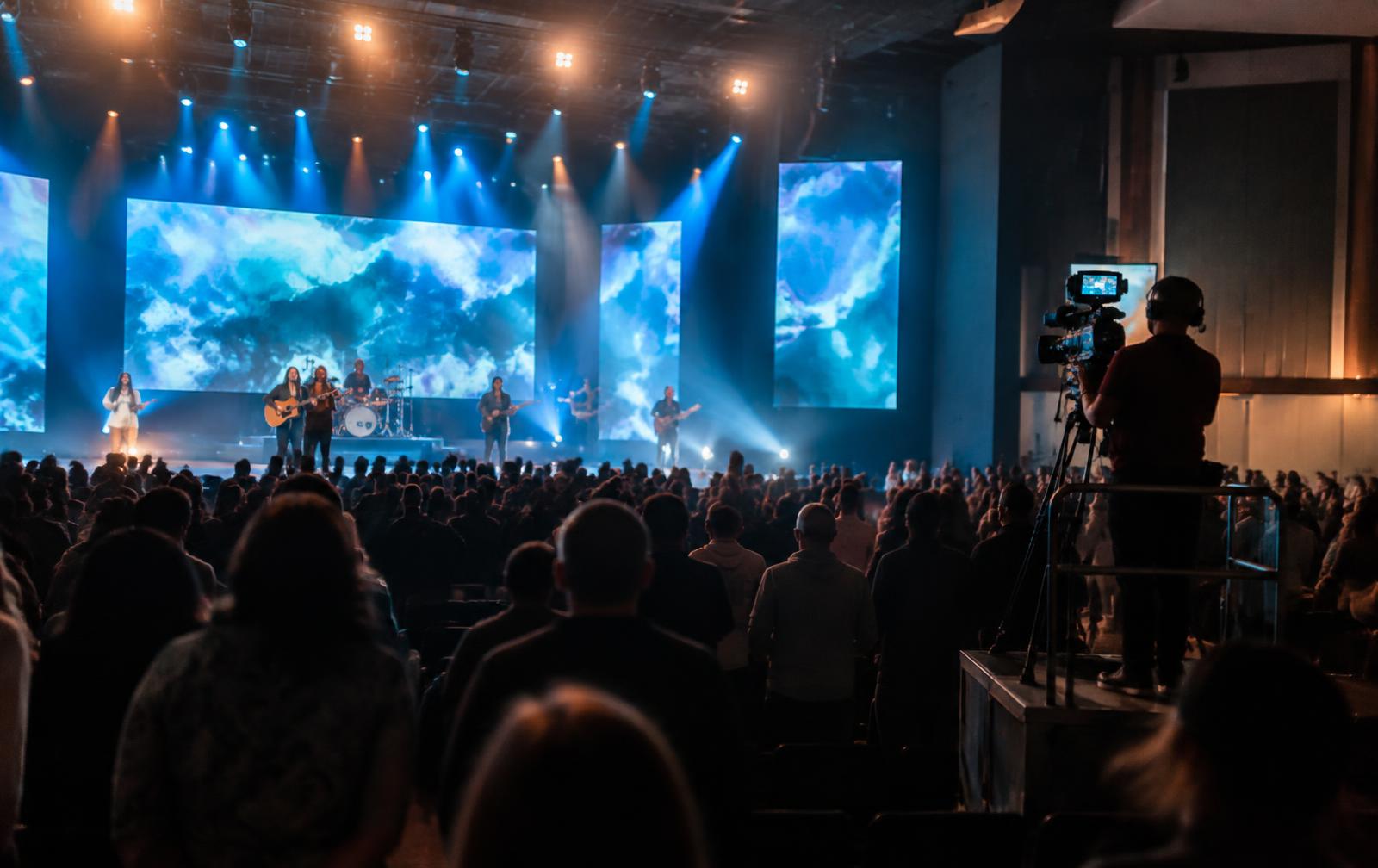 Worship service with band on a lit stage, LED screens and a camera operator on a riser