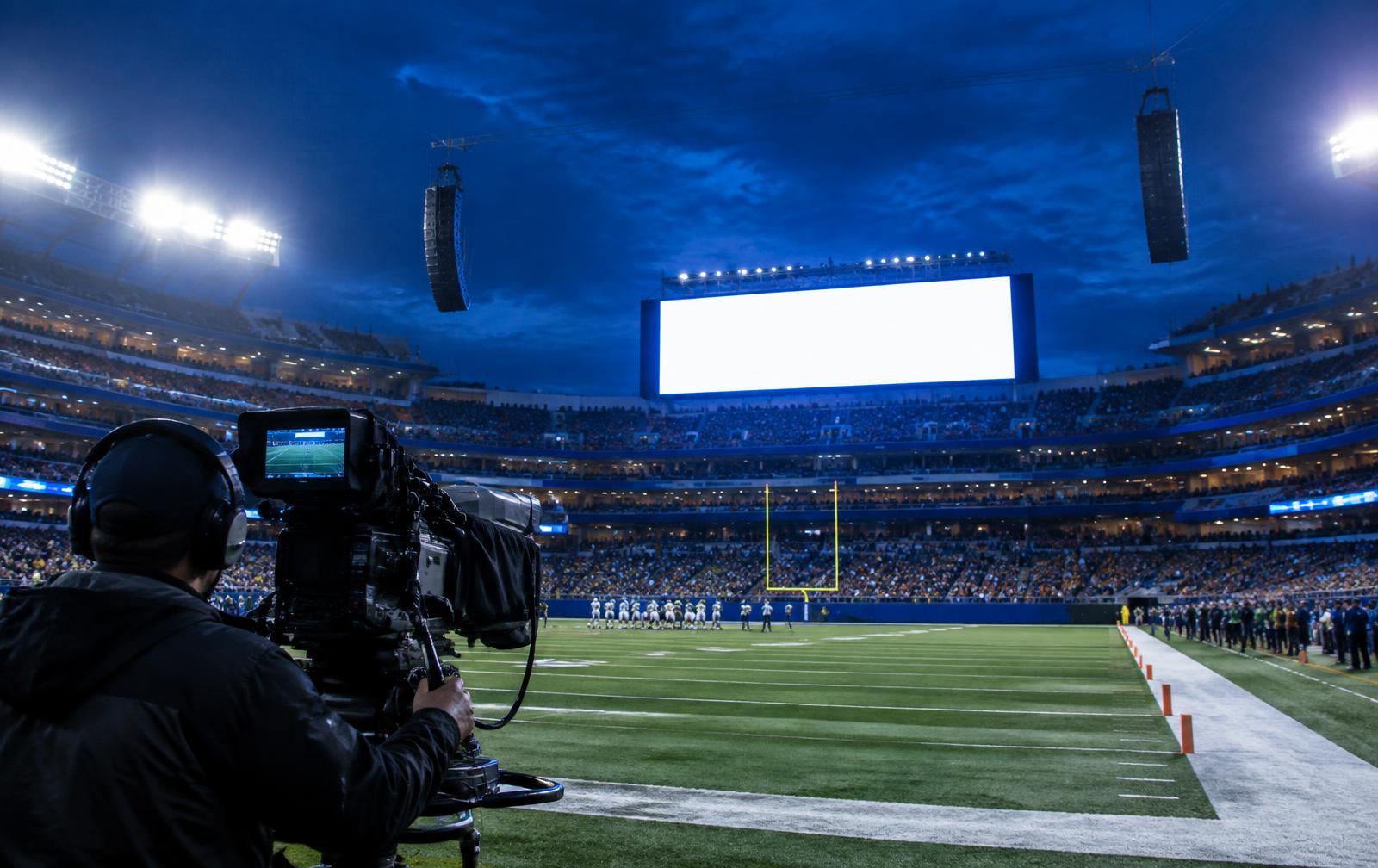 Sideline broadcast camera operator at a stadium with a video board and speaker clusters